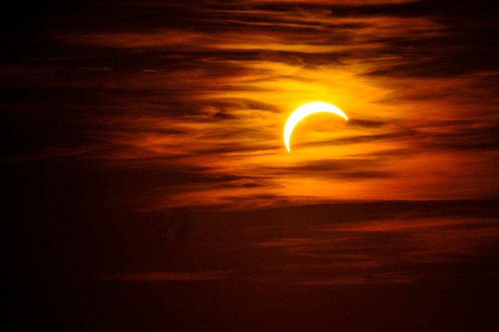 Thin crescent sun during the partial eclipse behind streaks of orange cloud