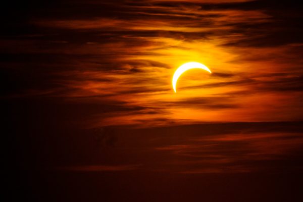 Thin crescent sun during the partial eclipse behind streaks of orange cloud